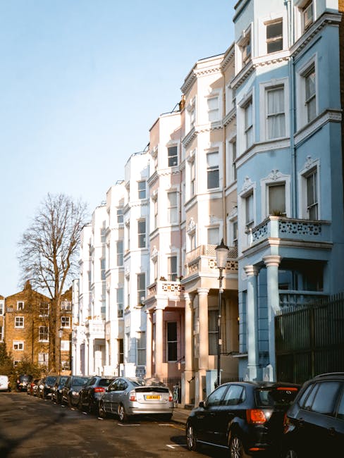 A line of multi-storey Victorian-style terraced houses with light blue and beige facades, decorative window mouldings, and small balconies. Cars are parked along the narrow street outside, with some t