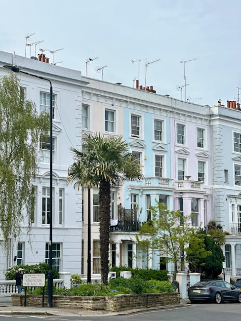 A row of white Victorian-style terraced houses with multiple windows, some with decorative molding, and small balconies at the front. A tall street lamp is positioned on the pavement next to a landscaped area with green shrubs, a palm tree, and a small tree. Several television antennas are visible on the rooftops, and parked cars are along the street in front of the properties. The scene reflects an urban residential area typical of Notting Hill, where home relocation and furniture transport services by Man With a Van Notting Hill often assist with moving houses in the neighbourhood. The lighting suggests daytime with a cloudy sky, and the overall environment is calm and orderly, suitable for smooth loading and unloading of belongings during a staircase or house removal involving packing and moving logistics.