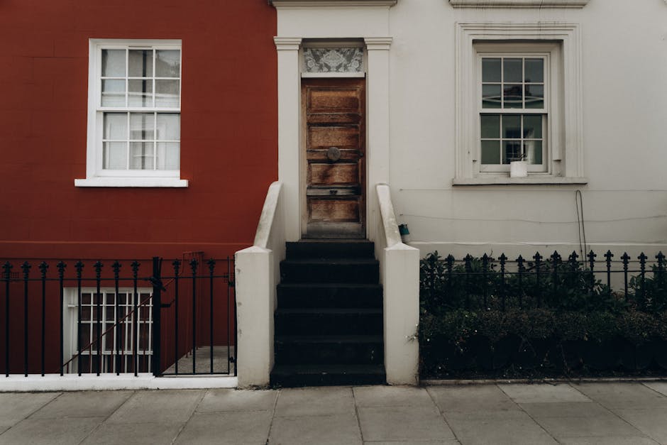 The image depicts the exterior of a residential property with a staircase leading up to an aged, wooden front door set between two adjacent buildings, one painted in a deep red and the other in white. The staircase is made of black material with white-painted concrete sides, and has a small handrail on each side. At the top of the stairs, the wooden door appears weathered, with visible horizontal panels, surrounded by decorative detailing on the lintel above. To the left, a white-framed window with six panes is visible on the red building, with a similar window on the white building to the right. The ground-level windows are partially obscured by a black metal fence with pointed finials, enclosing a small garden bed with low shrubs and plants. The scene is captured during daylight, with natural light illuminating the exterior walls and pavement in front. In the context of house removals, [COMPANY_NAME] might be involved in the logistics of interior furniture transport or staircase access during home relocation, as suggested by the arrangement of objects and access points in this setting.