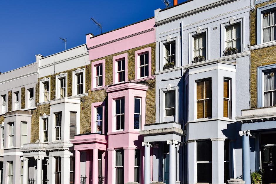 Photograph of a row of Victorian terraced houses in Notting Hill with colorful facades, primarily in shades of white, grey, and pastel pink. The buildings feature large sash windows, some with decorative window boxes and curtains. The photo is taken during daylight with a clear blue sky overhead. The houses are closely spaced, aligned along a pavement, with the facades showing architectural details such as cornices, bay windows, and brickwork. In the foreground, there are no visible moving activities or objects; however, this image visually represents the residential environment where house removals and packing services might be required. The image contextually aligns with home relocation and furniture transport services often conducted by companies like Man With a Van Notting Hill, highlighting the typical urban setting for end-of-tenancy clearance and moving logistics.