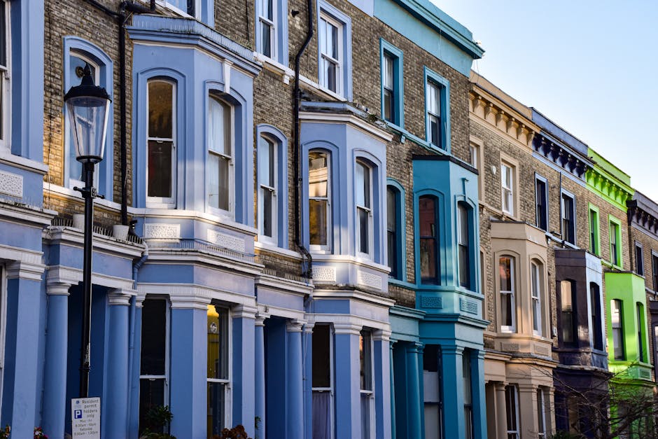 A row of three to four-storey terraced houses in Notting Hill with Victorian-style architecture, featuring large sash windows, decorative moldings, and painted facades in shades of light blue, cream, and vibrant green. The buildings are constructed from brick, with some sections covered in painted wood paneling, and are situated along a street with a parked van partially visible on the pavement. In the foreground, there is a black lamppost near the buildings. The image is taken during daytime with clear weather, highlighting the residential property exteriors, which are the focus of house removals and end-of-tenancy clearance services, such as those provided by Man With a Van Notting Hill.