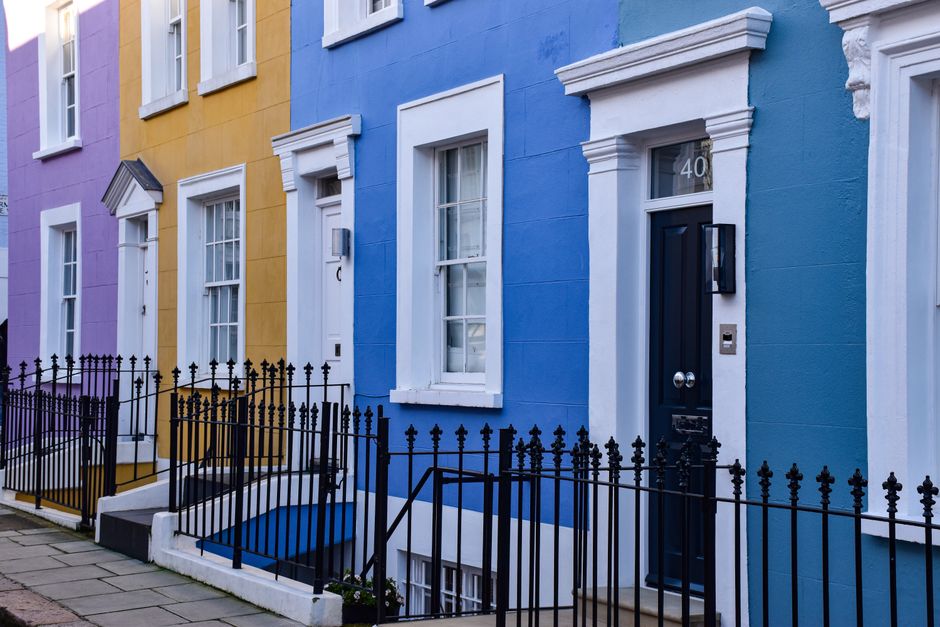 Photograph of a row of Victorian terraced houses in Notting Hill with colorful facades, primarily in shades of white, grey, and pastel pink. The buildings feature large sash windows, some with decorative window boxes and curtains. The photo is taken during daylight with a clear blue sky overhead. The houses are closely spaced, aligned along a pavement, with the facades showing architectural details such as cornices, bay windows, and brickwork. In the foreground, there are no visible moving activities or objects; however, this image visually represents the residential environment where house removals and packing services might be required. The image contextually aligns with home relocation and furniture transport services often conducted by companies like Man With a Van Notting Hill, highlighting the typical urban setting for end-of-tenancy clearance and moving logistics.