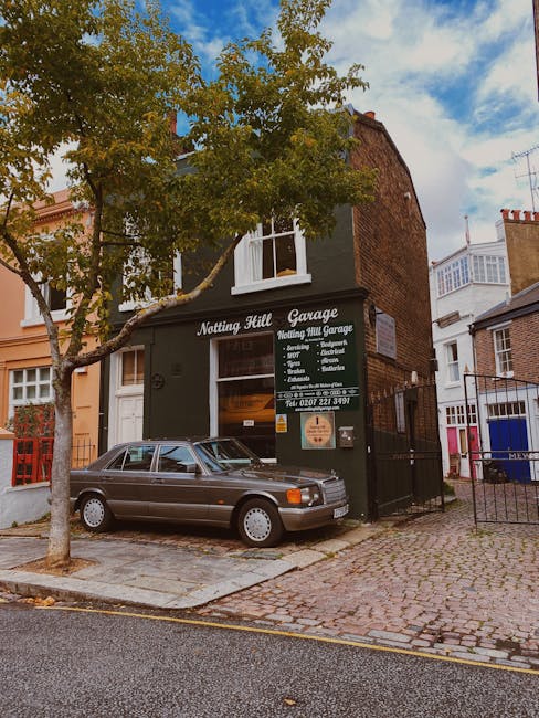 Exterior view of the Notting Hill Garage situated on a cobbled street, featuring a dark green storefront with white signage displaying 'Notting Hill Garage' and details of services such as removals, packaging, and storage. A vintage grey sedan is parked directly in front of the garage entrance, partially blocking the footpath. A large tree with green leaves partially obscures the upper portion of the building, with branches extending towards the shop window. The surrounding buildings are residential, with brick façades and bay windows, and other properties are visible to the right. The scene is illuminated by natural daylight with a partly cloudy sky overhead, indicating a typical day suitable for house relocations or furniture transportation involving planning and loading processes for home moves, as occasionally coordinated by companies like Man With a Van Notting Hill.