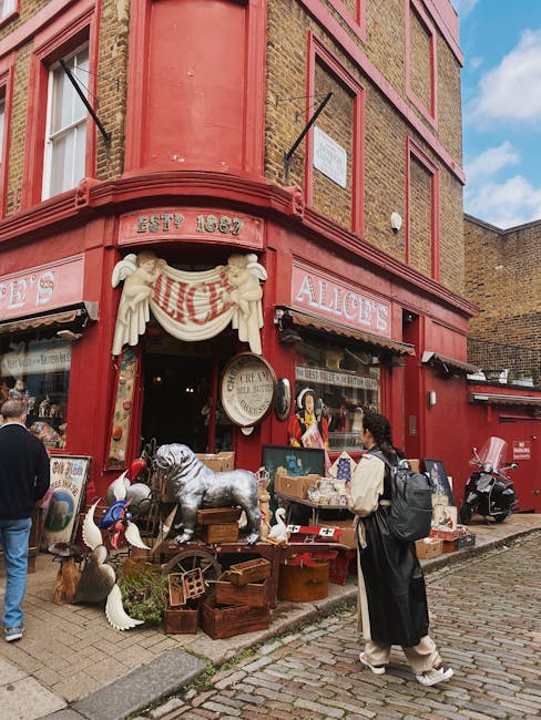 The exterior of a red brick building with a traditional pub frontage, decorated with a white draped banner and the establishment's name, 'Alice's,' displayed above the entrance. Outside, various vintage-style ornaments and collectibles, including a life-sized ceramic dog, wooden crates, and decorative figurines, are arranged on the pavement. Several people are visible, one woman with curly hair and a backpack is standing near the display, observing the items. The scene is set on a cobblestone street in Notting Hill, with clear daytime weather and a blue sky. The image captures the character of a busy area, potentially during a market or shopping event, aligned with local shopping or furniture transport services such as those provided by Man With a Van Notting Hill, which specializes in house removals and moving logistics.