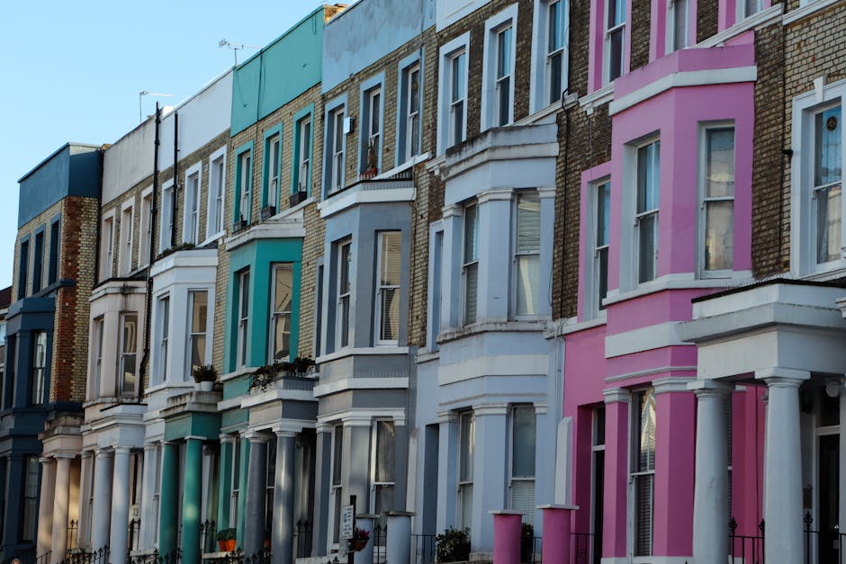 A row of Victorian-style terraced houses in Notting Hill, featuring colorful façades painted in shades of blue, white, and pink. The buildings have large front windows with white frames, some with window boxes containing plants. The houses display architectural details such as bay windows, decorative molding, and columns supporting small porches. The sky above is clear, and the street appears quiet. The image is taken during daylight, showcasing the vibrant exterior finishes. As part of a house removal process, [COMPANY_NAME] may be involved in transporting furniture or packing materials, which are not visible in this image, but the setting indicates an urban residential relocation environment.