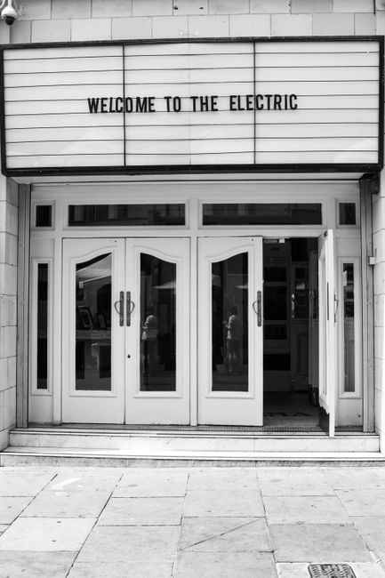 The black-and-white photograph shows the exterior of a building with a large, illuminated marquee sign reading 'WELCOME TO THE ELECTRIC' positioned above a set of white double doors with glass panels. The doors are framed by a white facade, and through the glass, reflections of people and interior elements are faintly visible. In front of the entrance, there is a paved sidewalk. Although the image does not depict furniture or moving activity, it gives a clear view of the building's entrance, which could be associated with a theatre or entertainment venue that may require professional house removals or relocation services by companies such as Man With a Van Notting Hill, especially when moving items into or out of this location.