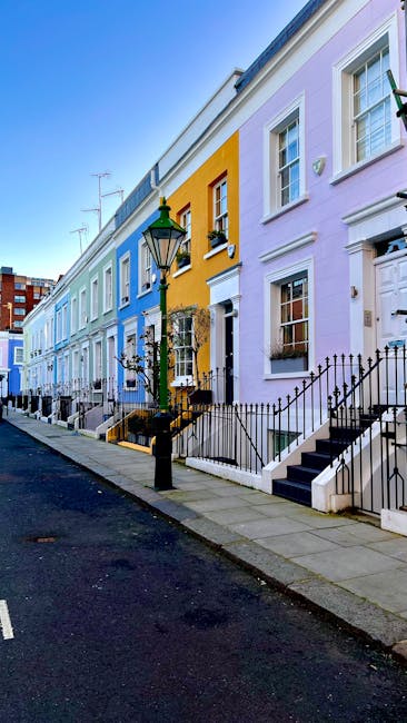 A row of Victorian-style terraced houses in Notting Hill, featuring colorful façades painted in shades of blue, white, and pink. The buildings have large front windows with white frames, some with window boxes containing plants. The houses display architectural details such as bay windows, decorative molding, and columns supporting small porches. The sky above is clear, and the street appears quiet. The image is taken during daylight, showcasing the vibrant exterior finishes. As part of a house removal process, [COMPANY_NAME] may be involved in transporting furniture or packing materials, which are not visible in this image, but the setting indicates an urban residential relocation environment.