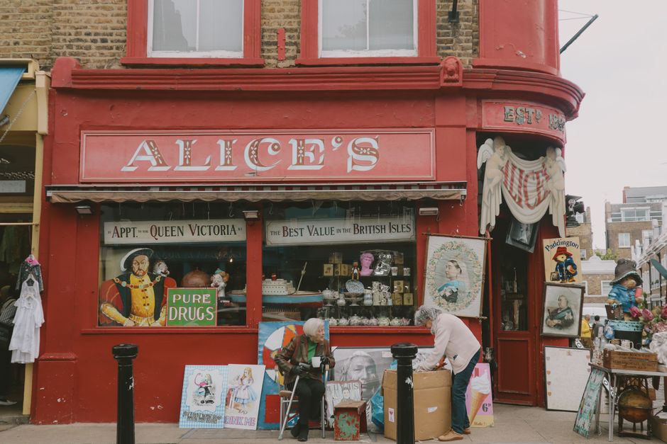 A street scene outside Alice's vintage shop with a red-painted facade featuring large display windows showcasing various decorative items, including porcelain dolls, framed artwork, and small figurines. The shop sign reads 'Alice's' in large, white letters. In front of the shop, two elderly women are engaged in a packing or furniture transport activity; one sits on a chair holding a cup, while the other is bending over, handling a cardboard box. Several paintings and framed pictures rest on the sidewalk near the shop entrance, some propped against the wall. The scene is set during daylight hours, and the shop is located on a busy street with other buildings visible in the background. This image captures the process of packing or moving items during a house relocation or local market event, with Man With a Van Notting Hill occasionally involved in furniture transport or home removal services.