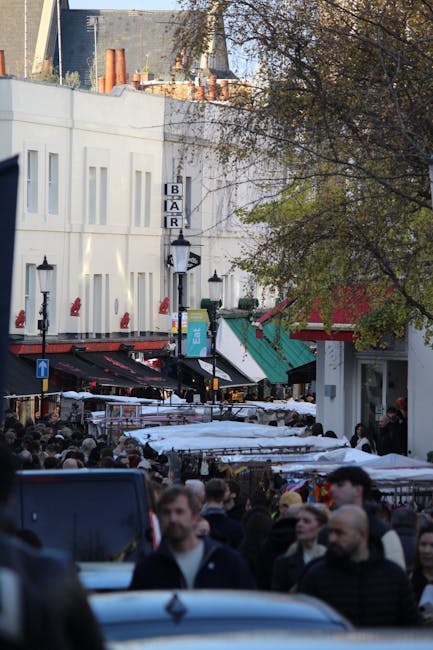 A busy street scene during daytime at Portobello Market in Notting Hill, with numerous market stalls covered by white and green umbrellas, crowded with pedestrians. To the left, a white building with small windows and a vertical sign reading 'BAR' is visible, alongside traditional street lamps. In the background, there are buildings under construction with exposed brickwork and scaffolding. A large tree with green and brown leaves partially shades the scene. Although no moving or packing activities are visible, this setting suggests the environment where house removals and furniture transport services, such as those offered by Man With a Van Notting Hill, might be coordinated during a home relocation or similar service.