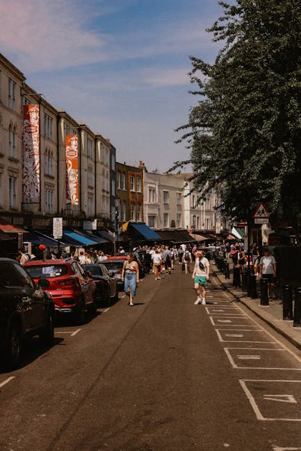 A busy street scene in Notting Hill during daytime, with pedestrians walking along the pavement and carrying boxes, furniture, and packing materials as part of a home relocation process. On the left side, parked cars line the curb, and above, multi-storey terraced houses with large banners hanging from some windows are visible. The houses feature a mix of brick and painted facades, with awnings over shop entrances at ground level. A large green tree extends across the right side of the image, providing partial shade. In the background, people are loading household items into a van parked on the street, with some objects wrapped in protective packaging such as plastic and blankets. The environment appears active, with movement indicating an ongoing furniture transport or packing and moving operation by a professional removals service, likely Man With a Van Notting Hill, in preparation for a house move.