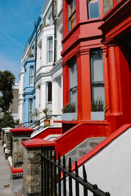 A row of brightly coloured terraced houses on a residential street, with the facades painted in shades of blue, white, and red. The houses feature bay windows and small front gardens enclosed by black wrought-iron fences. Concrete steps with red-painted sides lead up to each entrance, some of which are partially visible. In the foreground, the entrance to the red house has potted plants on a small landing, while a man with a van from Man With a Van Notting Hill is involved in a home relocation process, preparing for furniture transport. The scene is lit by daylight, with a clear blue sky overhead, and the surroundings include a few trees and urban street elements, illustrating an environment suitable for packing and moving services.