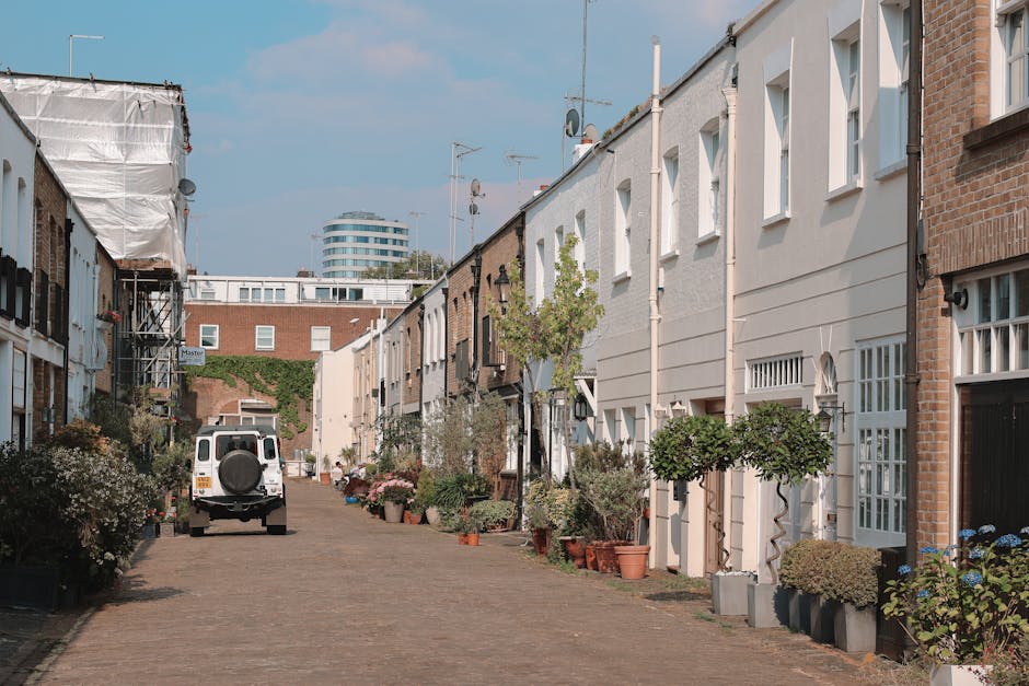 A narrow residential street in Notting Hill lined with multi-storey houses featuring brick and white-painted facades, with potted plants and small trees decorating the front entrances. In the foreground, a white van from Man With a Van Notting Hill is parked on the cobbled pavement, positioned near the open doorway of one of the houses, indicating the loading process involved in home relocation. The van's rear doors are open, and cardboard boxes, wrapped furniture, and moving blankets are visible inside, reflecting packing and furniture transport activities. A hand truck or trolley may be in use to assist with lifting or carrying items. The street has a slight downward slope, with additional houses and an urban multi-storey building visible in the background, under a partly cloudy sky. The scene captures the typical logistics of household removals, emphasizing careful packing, loading, and transport performed by professional movers like Man With a Van Notting Hill.