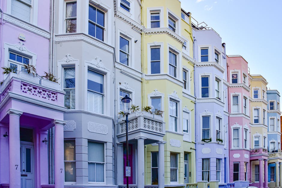 A narrow residential street in Notting Hill lined with multi-storey houses featuring brick and white-painted facades, with potted plants and small trees decorating the front entrances. In the foreground, a white van from Man With a Van Notting Hill is parked on the cobbled pavement, positioned near the open doorway of one of the houses, indicating the loading process involved in home relocation. The van's rear doors are open, and cardboard boxes, wrapped furniture, and moving blankets are visible inside, reflecting packing and furniture transport activities. A hand truck or trolley may be in use to assist with lifting or carrying items. The street has a slight downward slope, with additional houses and an urban multi-storey building visible in the background, under a partly cloudy sky. The scene captures the typical logistics of household removals, emphasizing careful packing, loading, and transport performed by professional movers like Man With a Van Notting Hill.
