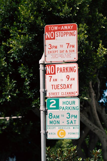 A vertical street signpost equipped with multiple parking restriction notices is situated outdoors amidst green foliage and tall trees. The top sign is red and white, indicating a tow-away zone with no stopping allowed from 3 pm to 7 pm, except on Saturdays and Sundays for towed cars, and includes a contact phone number for further details. Below, a second sign, also in red and white, specifies no parking from 7 am to 9 am on Tuesdays for street cleaning purposes. The third sign is green and white, designating a two-hour parking limit from 8 am to 3 pm, Monday through Saturday, for vehicles with a valid C permit. The signpost is likely positioned near a residential area in Notting Hill, supporting residents and visitors with clear parking regulations related to house removals or relocation services such as those provided by Man With a Van Notting Hill, which specializes in removals and furniture transport. The signage is clearly visible under natural light, with the trees providing a scenic backdrop, and the signs appear well-maintained and easily legible for drivers and pedestrians alike.