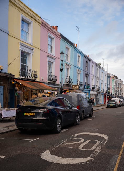 A street scene in Notting Hill featuring a row of colorful terraced houses painted in pastel shades of yellow, pink, blue, and white, with decorative wrought iron balconies and window frames. The pavement is occupied by parked vehicles including a black car and a grey van positioned near the curb, adjacent to a parking zone marked with a white '20' speed limit sign painted on the road. In front of the houses, there are small storefronts with awnings, visible signage, and outdoor displays, indicating commercial activity. The image appears to be taken during daylight hours with clear weather and a partly cloudy sky. This setting reflects the typical environment where house removals and furniture transport may take place, with residents and removal companies managing packing, loading furniture into vans, and navigating parking regulations, such as W11 parking permits, to avoid fines. The presence of a Man With a Van Notting Hill vehicle suggests professional moving services involved in home relocation or furniture transport in this picturesque neighbourhood.